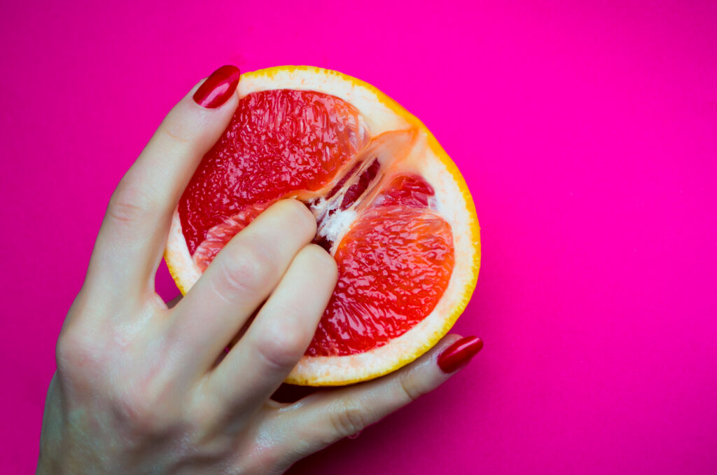A two fingers pressing into a grapefruit, symbolizing female sexuality against a vibrant pink background