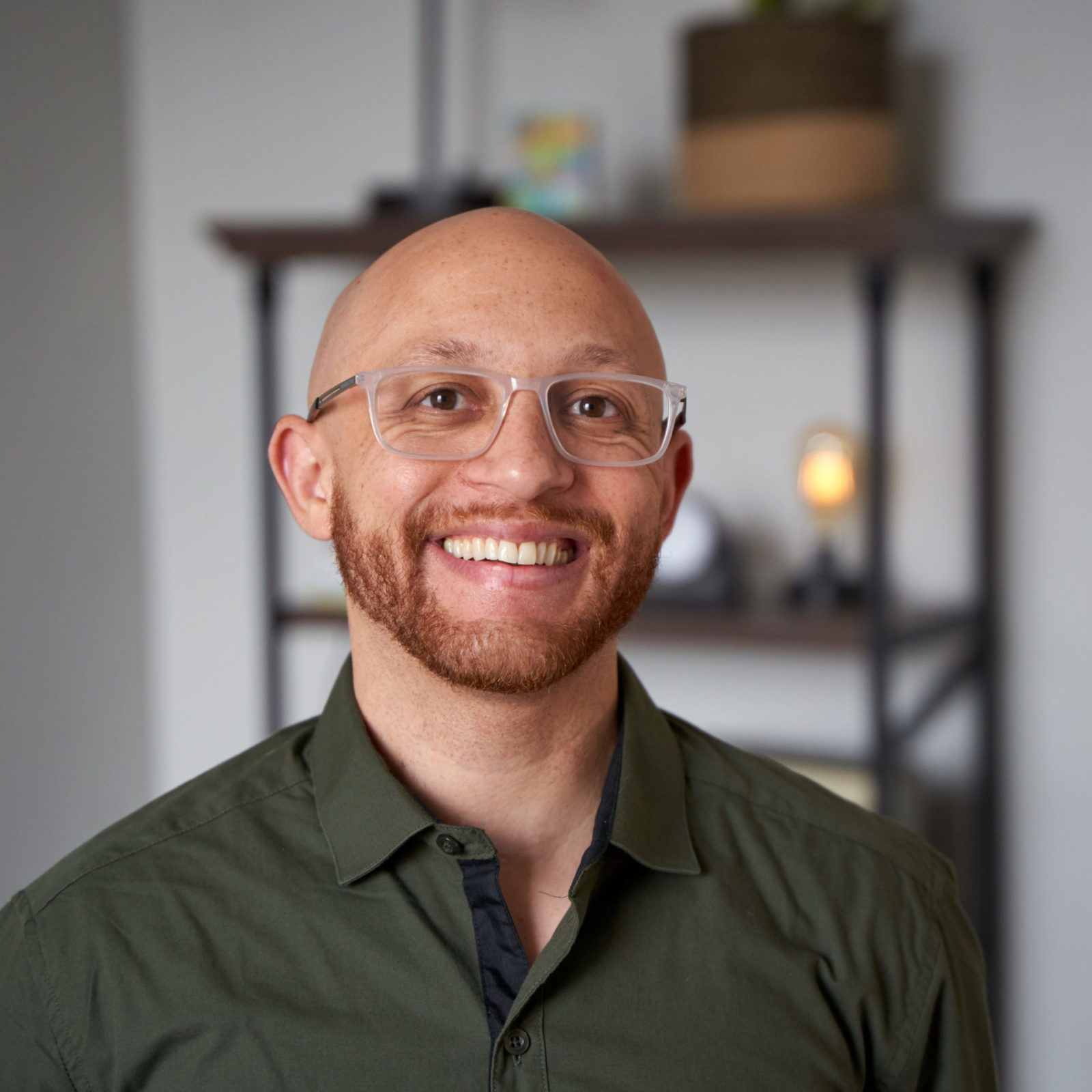Caleb Dunn, therapist in Philadelphia with red facial hair, shaved head and brown eyes. Caleb Dunn is wearing a dark green button up shirt and clear glasses while looking at the camera smiling in an office at The Better You Institute with a shelf and light in the background.
