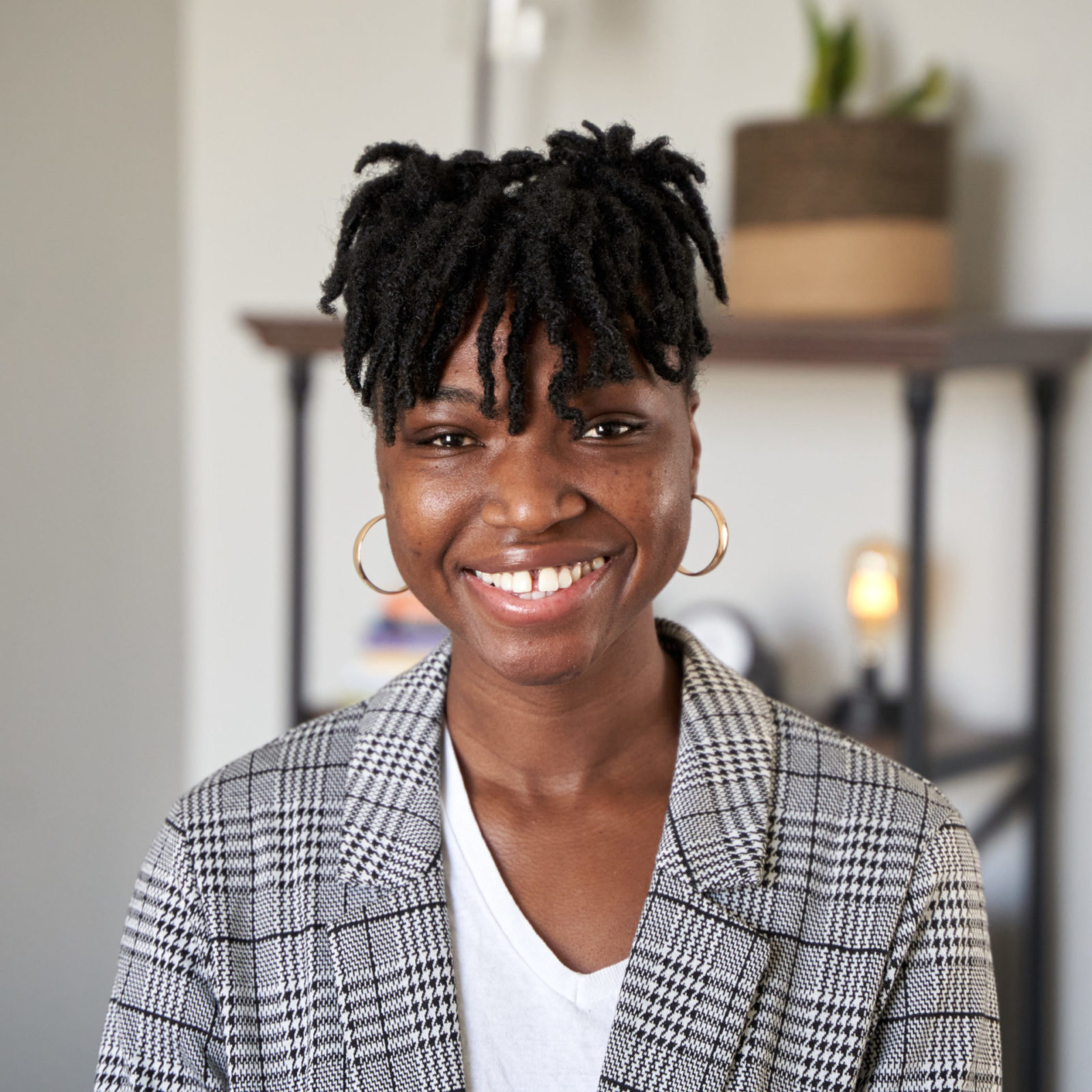 Hilda “Madequor” Tetteh-Ocloo, therapist in Philadelphia with brown dreadlock hair and brown eyes. Hilda is wearing golden hoop earrings with white shirt and a geometric pattern blazer. Hilda is looking at the camera smiling in an office at The Better You Institute with a shelf and light in the background.
