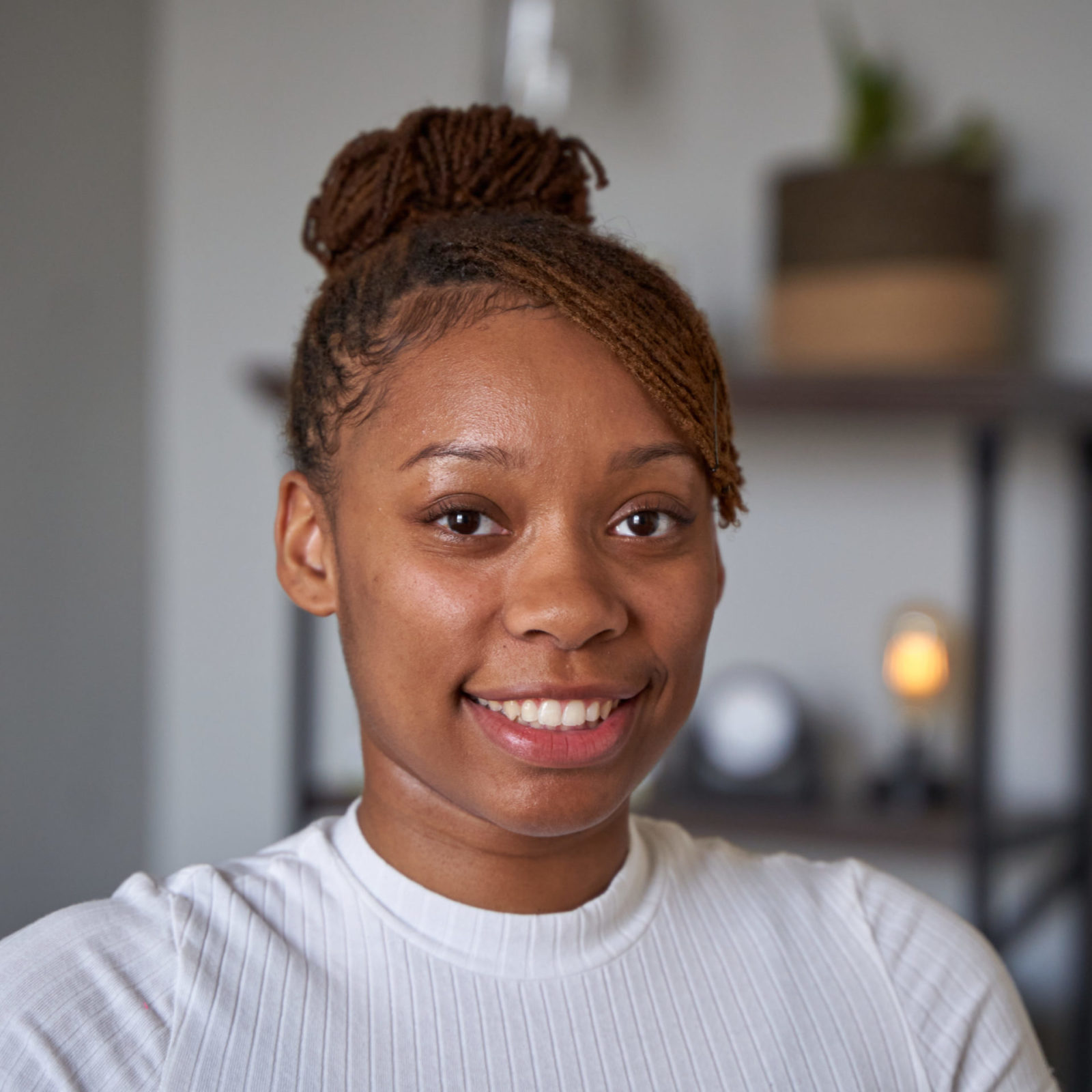 Sabree Wright, therapist in Philadelphia with brown hair and brown eyes. Sabree is wearing white shirt with her hair in a bun. Sabree is looking at the camera smiling in an office at The Better You Institute with a shelf and light in the background.