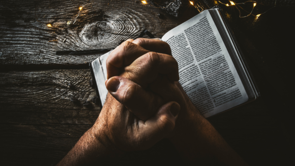Hands clasped in prayer resting on an open Bible on a wooden table with soft string lights in the background