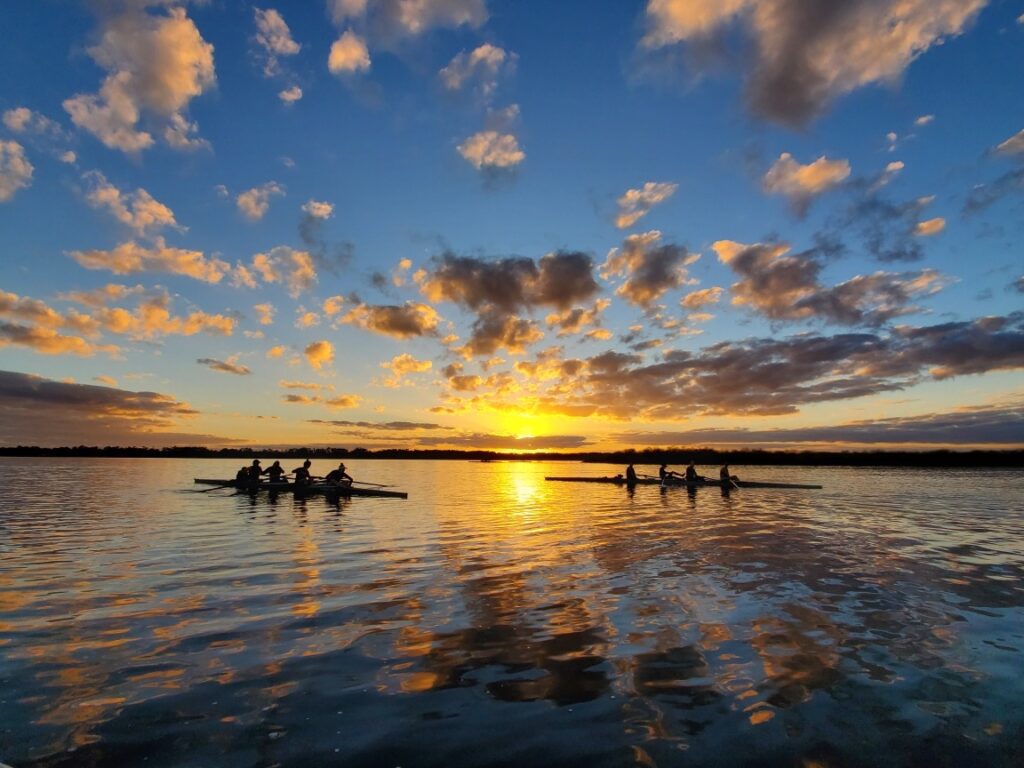 Rowers gliding across a calm lake at sunset, with golden hues reflecting off the water and scattered clouds in the sky
