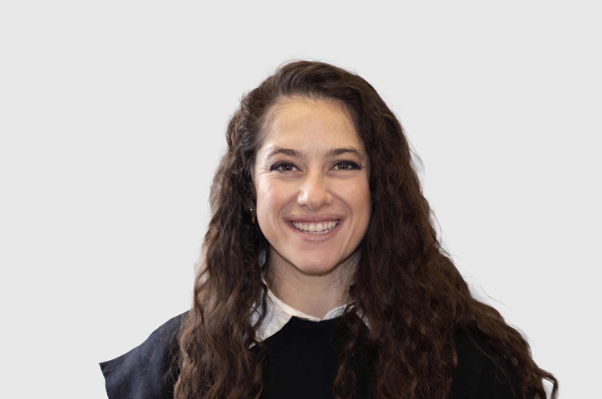 Hannah Goldstein, Psy.D., with long brown curly hair, smiling with teeth, wearing a black top with a white collar shirt underneath