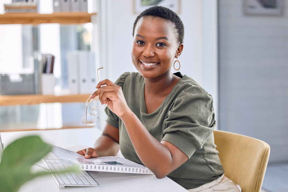 Smiling woman holding glasses while working at a desk with laptop and papers