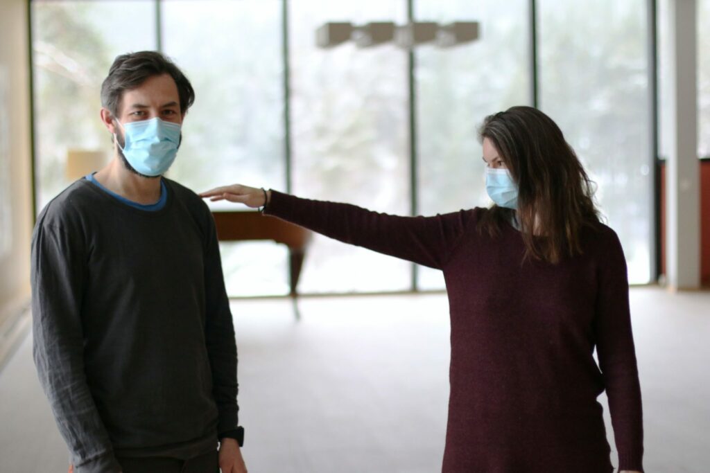 Two people wearing face masks standing indoors, maintaining distance as the woman extends her arm to touch the man’s shoulder