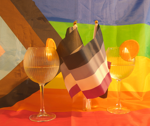Two glasses of drinks with orange slices sit in front of pride flags, including an asexual pride flag, symbolizing celebration and LGBTQ+ pride