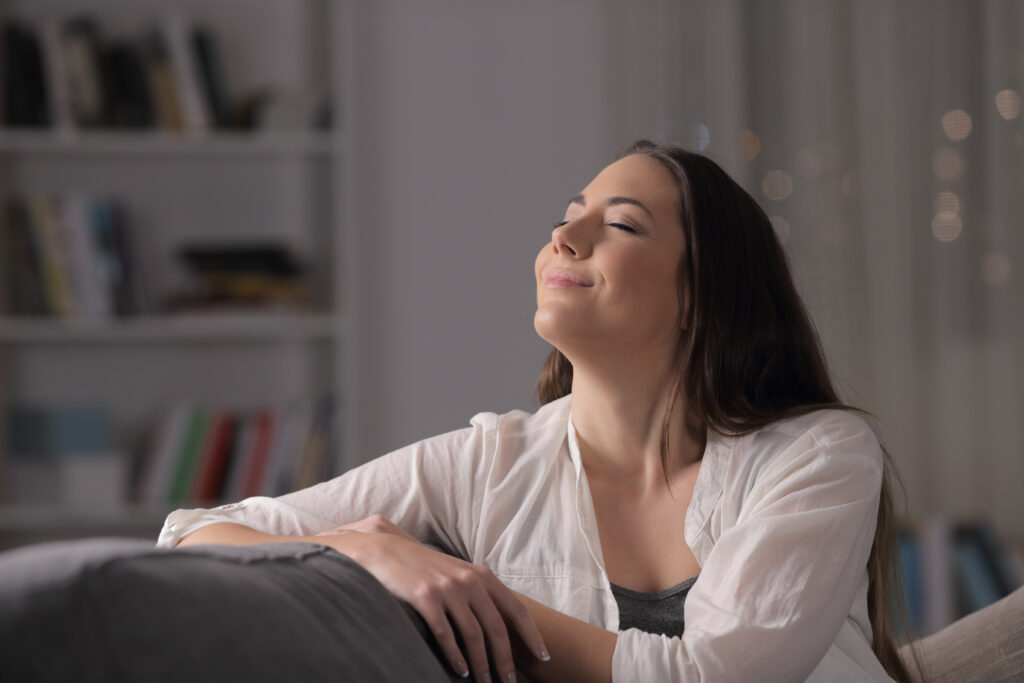 A woman sitting on a couch with her eyes closed and a relaxed smile, symbolizing peace, contentment, and emotional well-being
