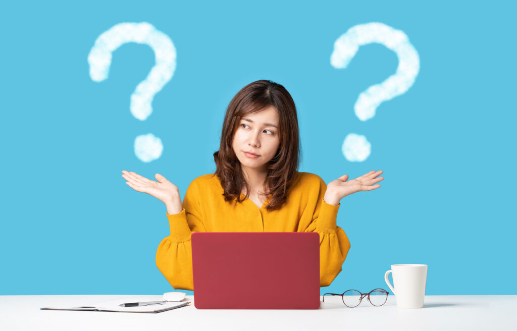 A woman shrugging at a desk with a laptop, surrounded by question marks, representing confusion or indecision