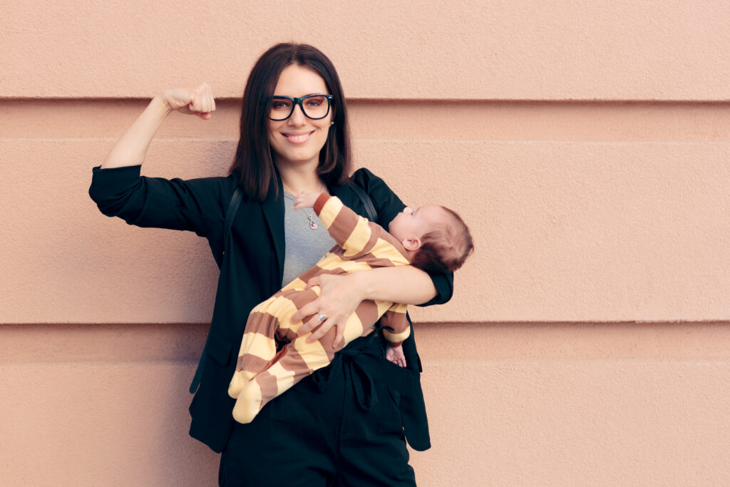 A smiling woman holds a baby while flexing her arm, symbolizing strength and resilience in motherhood