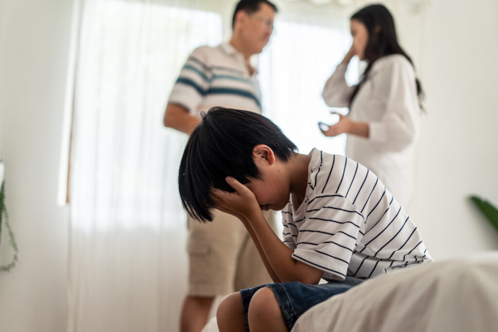 A child sitting on a bed, covering his face while parents argue in the background