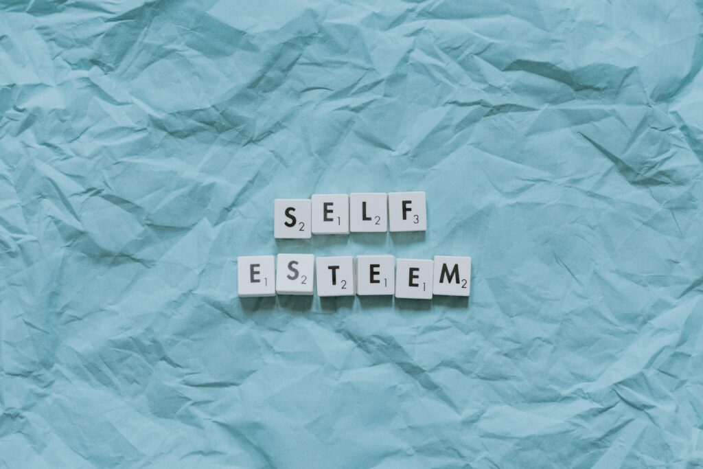 Scrabble tiles spelling "SELF ESTEEM" arranged on a crumpled blue paper background, symbolizing confidence, self-worth, and personal growth